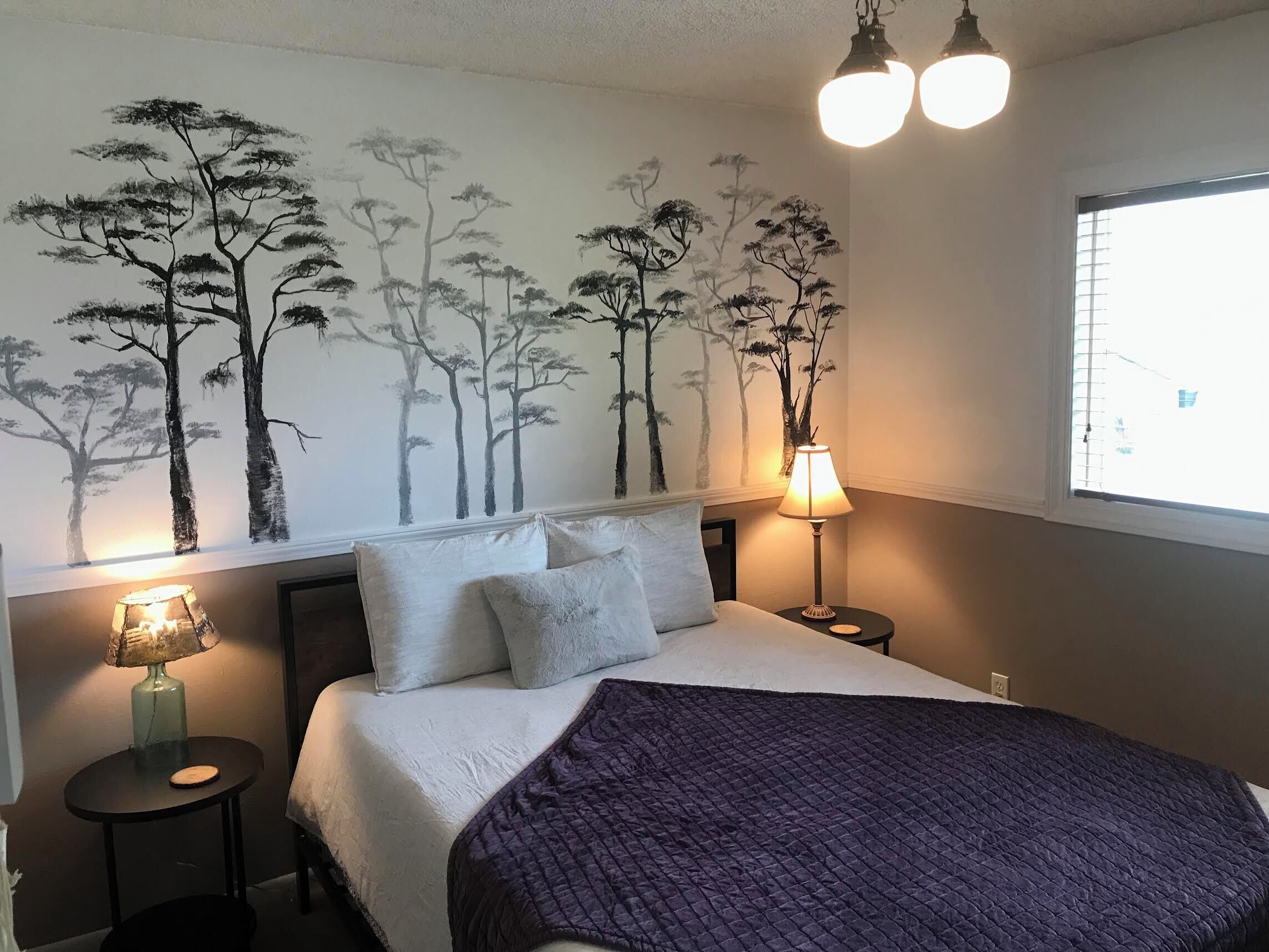 A bedroom with a bed, pillows, a blanket, a wall-mounted lamp, and a ceiling light fixture.