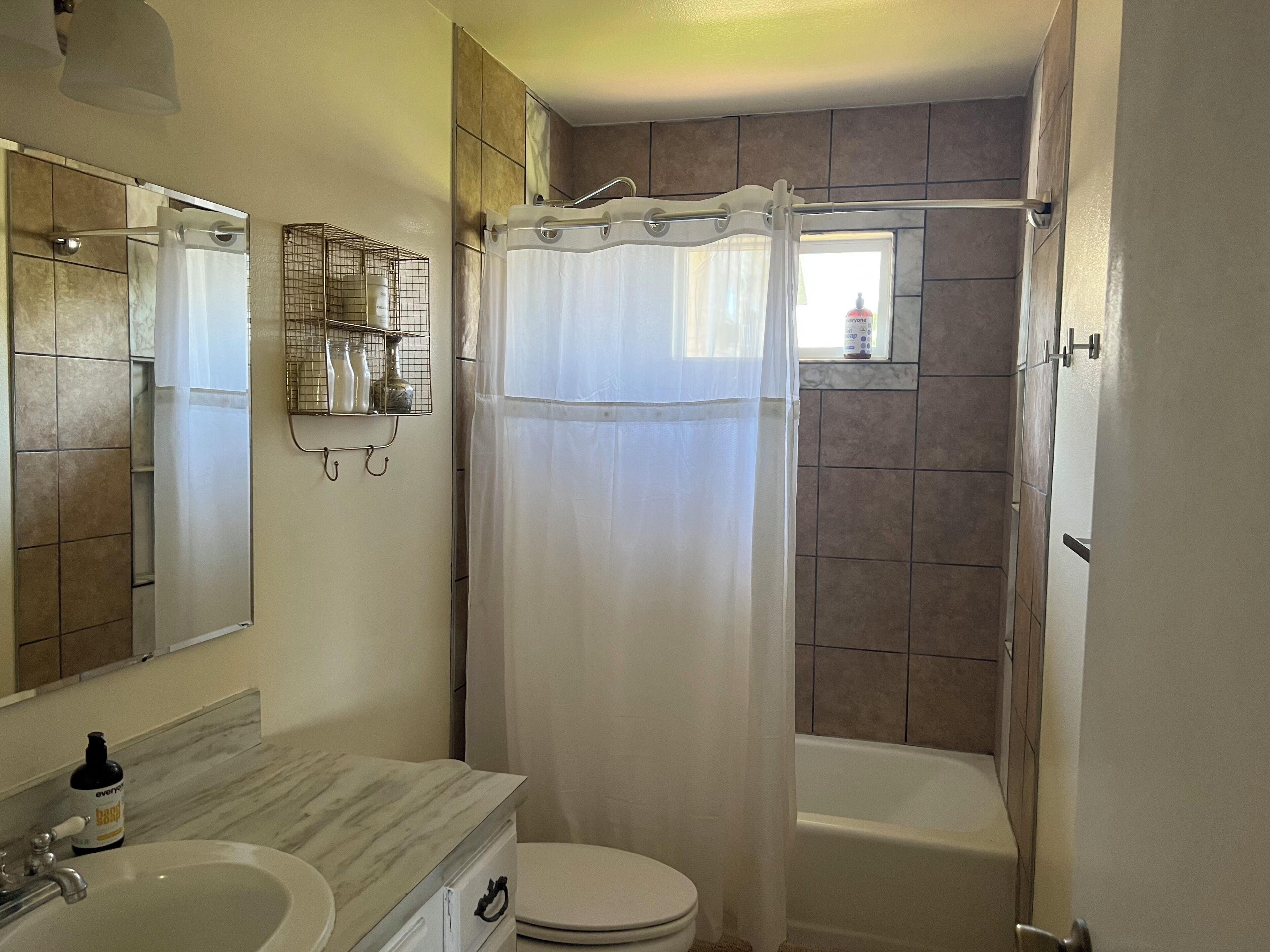 Een badkamer met toilet, wastafel en bad. Er is een douchegordijn, een plank met toiletartikelen, en een raam.