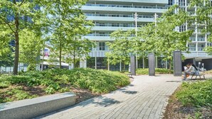 Un sendero pavimentado con vegetación, bancos y un edificio moderno de varios pisos al fondo.