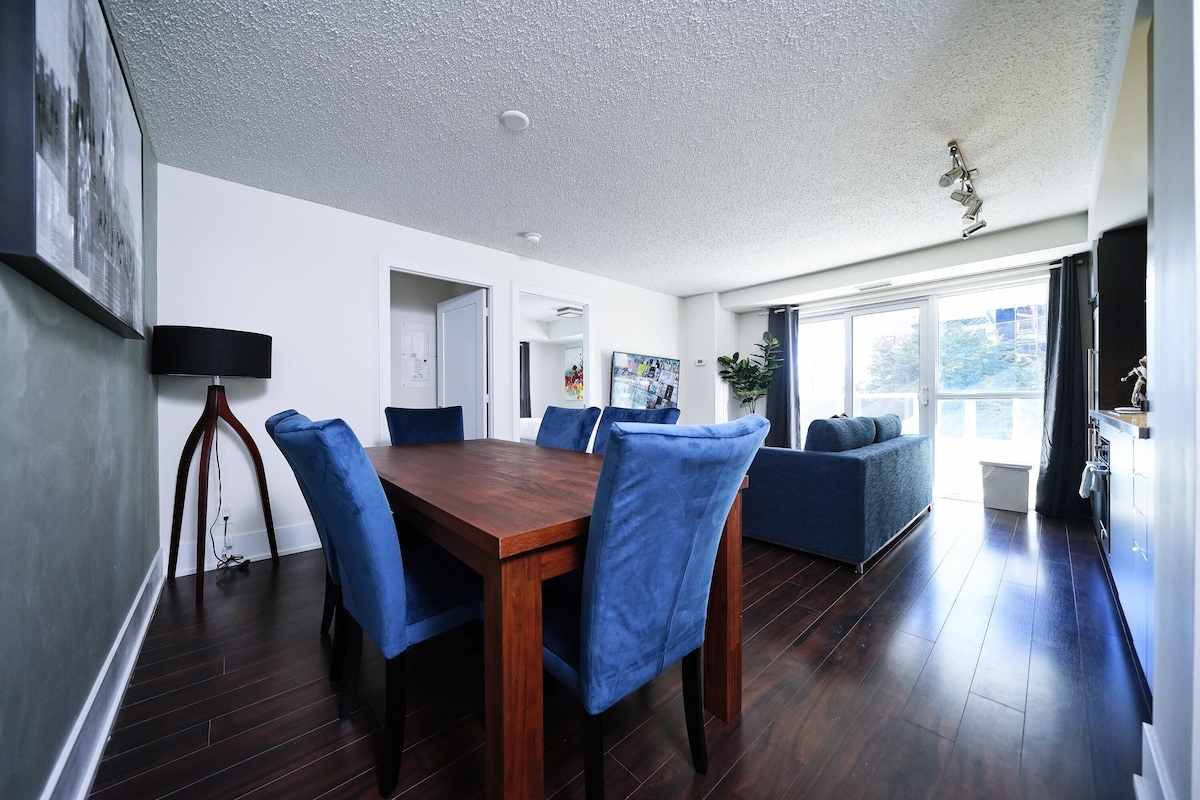 A modern dining area with a wooden table and blue chairs, a sofa, and a large window.