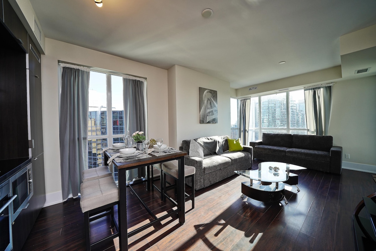 A modern living room with a dining area, dark wooden flooring, a grey sofa, a coffee table, and large windows with curtains.