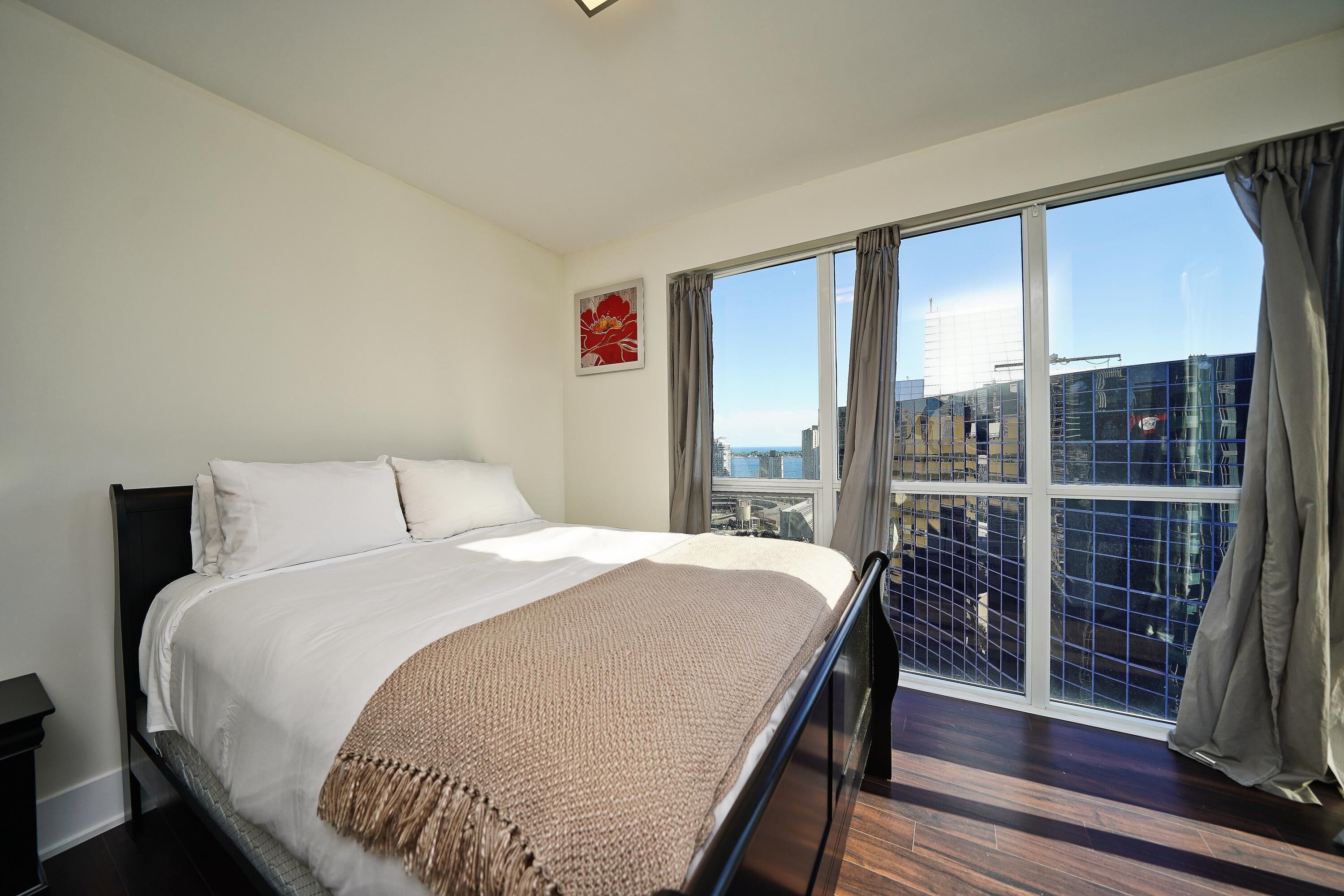 Une chambre d’hôtel avec un grand lit, une vue sur la ville grâce à de grandes fenêtres du sol au plafond, et un panorama urbain visible à l’extérieur.