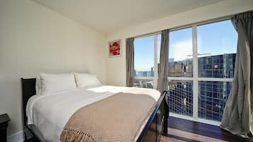 Une chambre d’hôtel avec un grand lit, une vue sur la ville grâce à de grandes fenêtres du sol au plafond, et un panorama urbain visible à l’extérieur.