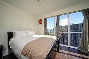 A hotel room with a large bed, a view of the city through floor-to-ceiling windows, and a cityscape visible outside. - StayToronto - The Atmos Suites (Toronto)