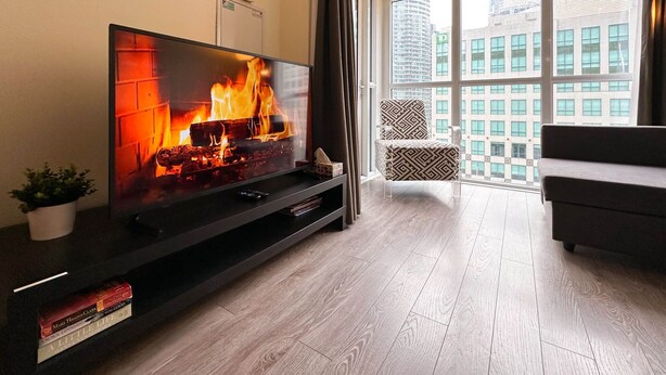 Una sala de estar moderna con un televisor grande que muestra una chimenea, un sofá y vistas de un paisaje urbano a través de amplios ventanales.