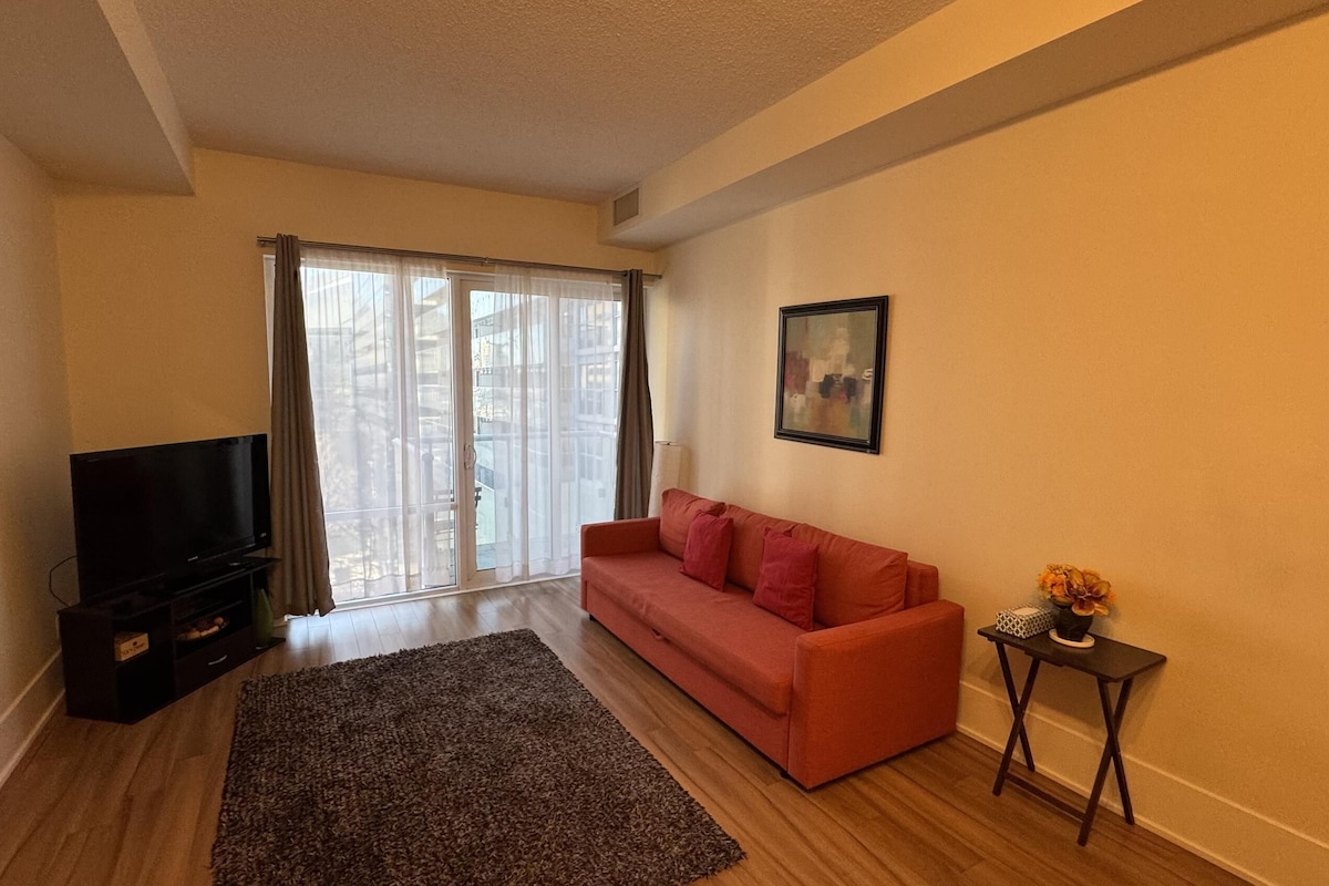 A living room with a sofa, a TV, a small table with a vase, and a large window with curtains.