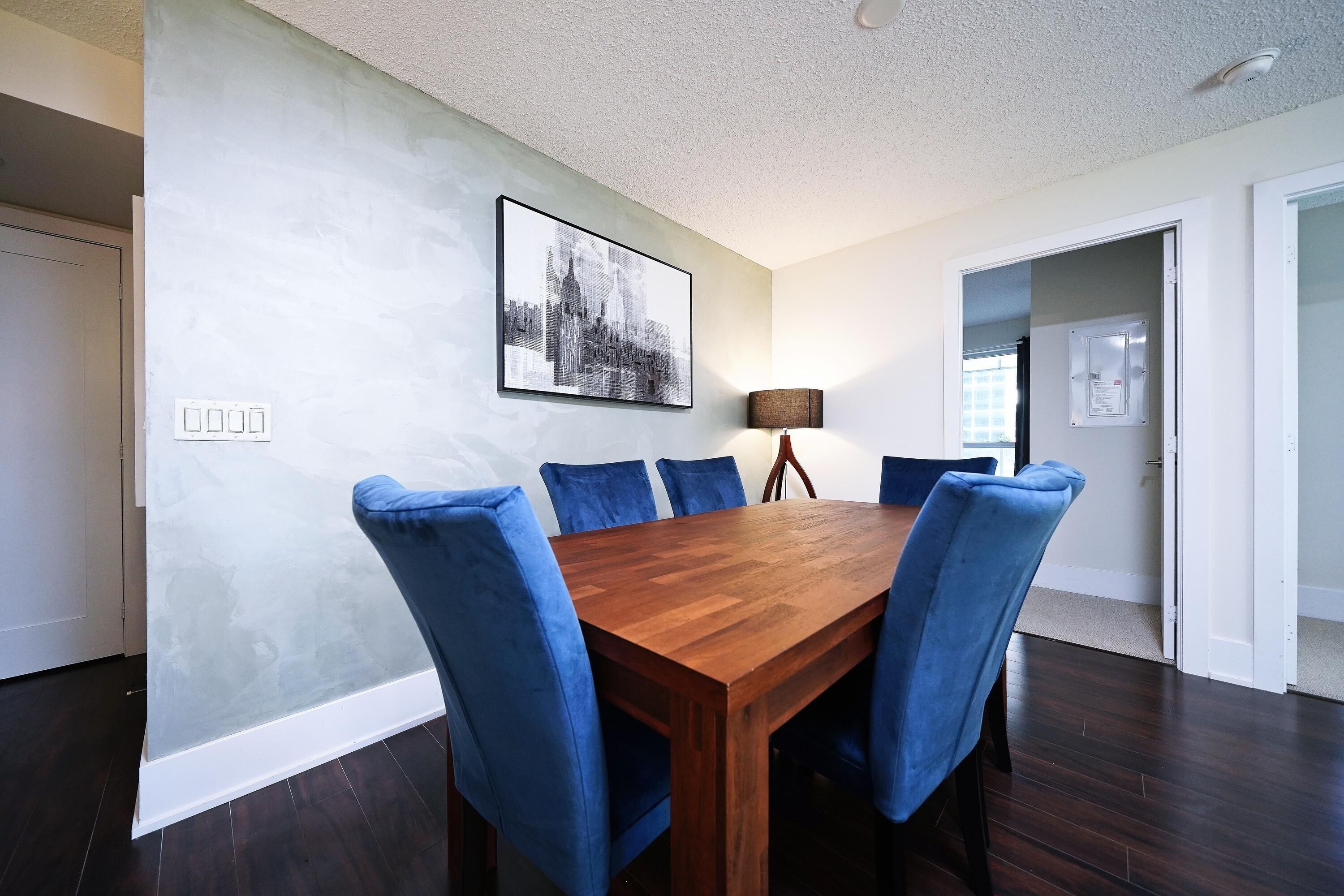 A dining area with a wooden table and blue chairs, a cityscape artwork on the wall, and a door leading to another room.