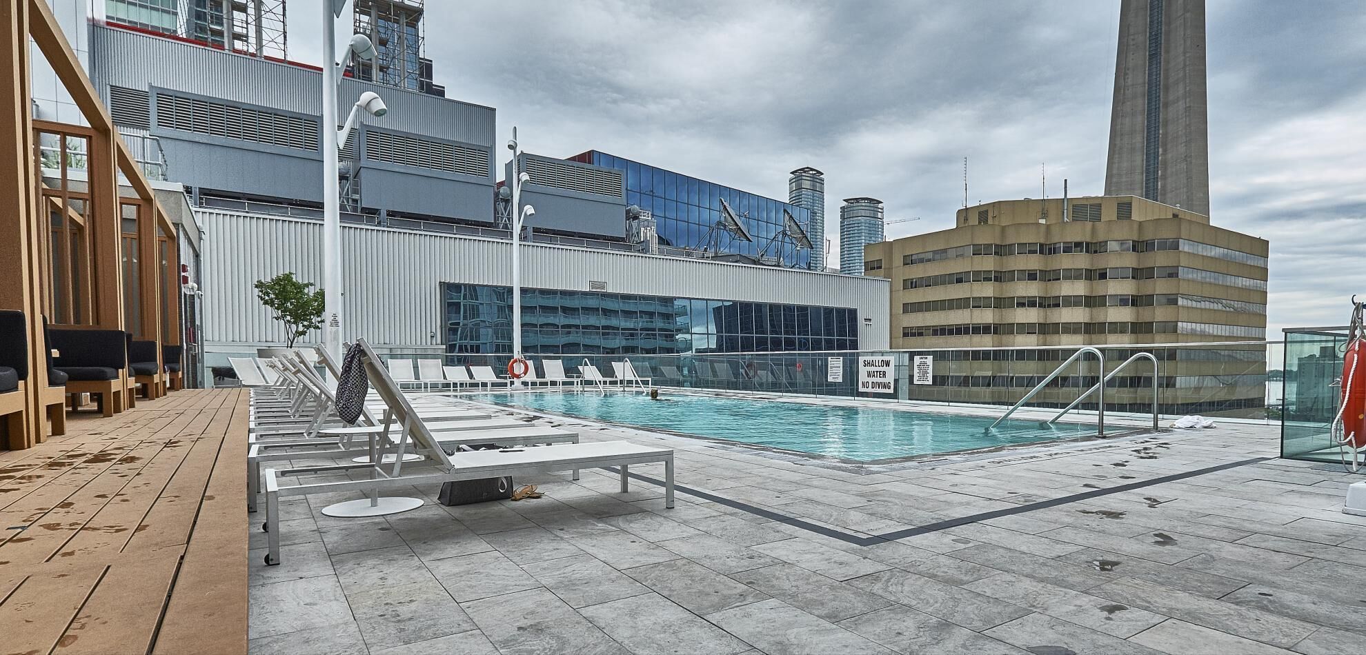 A rooftop pool with lounge chairs, a shallow water warning sign, and modern buildings in the background.