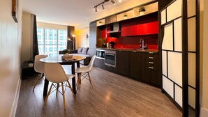 Una cocina moderna con armarios rojos, un comedor con una mesa redonda y sillas, y vistas de un edificio a través de amplios ventanales.
