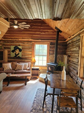 A log cabin interior with a wooden ceiling, a sofa, a dining table, and a fireplace. - cozy mountain retreat with wood stove and hot tub (Mena)