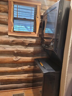A log cabin interior with a built-in washer and dryer, a window with blinds, and a wooden floor.