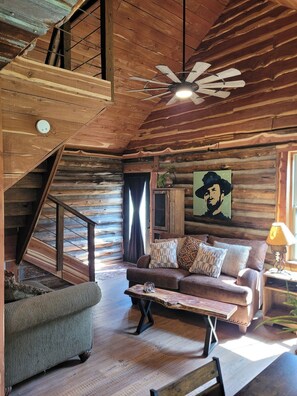A log cabin interior with a staircase, a ceiling fan, a sofa with patterned cushions, and a painting on the wall.
