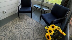 Una habitación con un ventanal grande, dos sillas, una mesita, un tapete y un florero con girasoles.