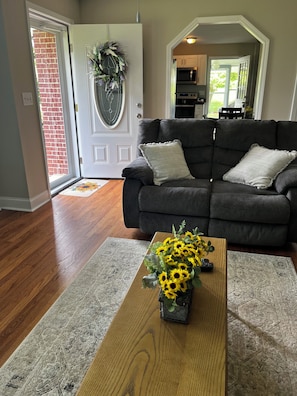 Un salon avec un canapé gris foncé, une table basse en bois ornée de tournesols, un tapis gris à motifs et une porte blanche décorée d’une guirlande.