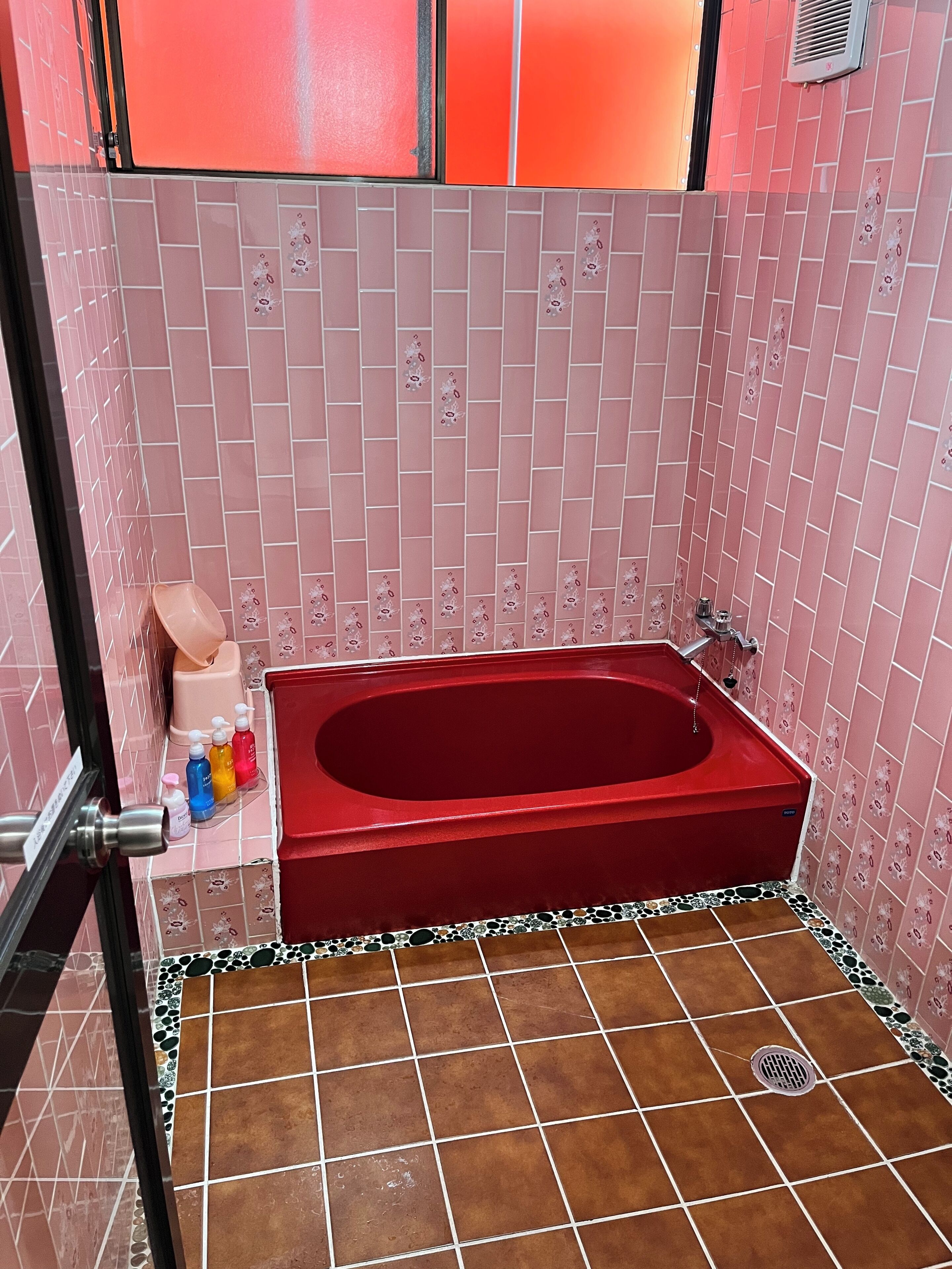 Een badkamer met een rood bad, roze betegelde wanden en een betegelde vloer met decoratieve rand.