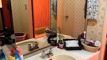 A bathroom with two sinks, a mirror, and various toiletries. There is a door with a floral pattern, a potted plant, and slippers on the floor.