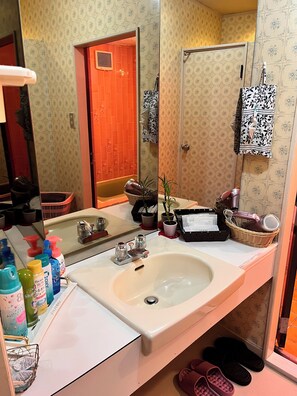 Een badkamer met twee wastafels, een spiegel en diverse toiletartikelen. Er is een deur met een bloemendesign, een potplant en slippers op de vloer.