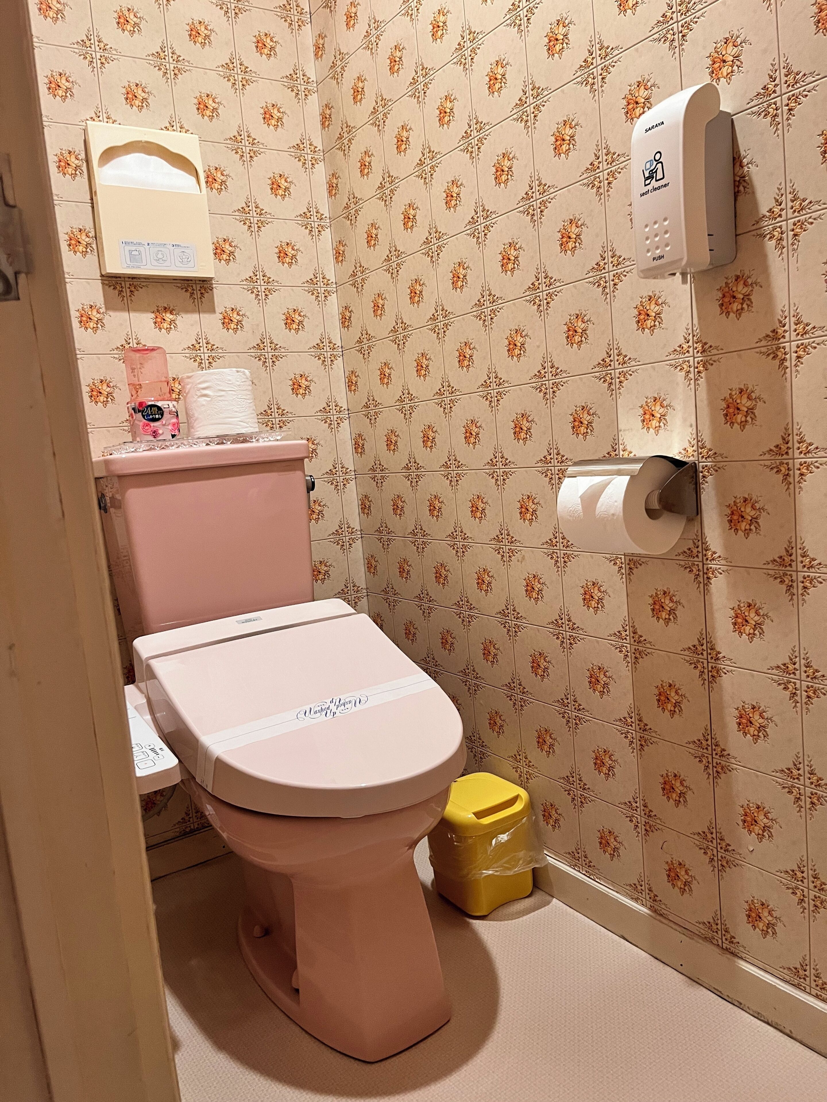 A bathroom with a pink toilet, a toilet paper holder, a trash bin, and a seat cleaner device on the wall.