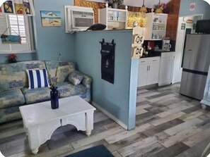 Uma sala de estar com um sofá, uma mesa de centro branca e uma área de cozinha com armários e uma geladeira.