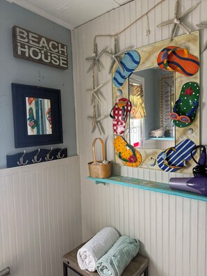 Um banheiro com espelho, porta-toalhas, prateleira com objetos de praia e uma placa que diz 'BEACH HOUSE'.