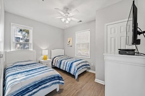 A bedroom with two beds, a ceiling fan, a television on a stand, and a window with blinds. - Ocean Keyes 3BR First Floor Condo - North Myrtle Beach (North Myrtle Beach)