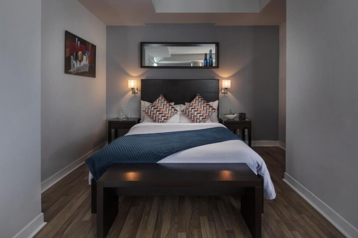 A modern bedroom with a dark headboard, two bedside tables with lamps, a large bed with patterned pillows, and a painting on the wall.