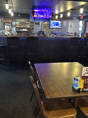 A bar area with wooden chairs and a bar counter, a television mounted above the bar, and a table with a bottle of mustard and a yellow condiment bottle.