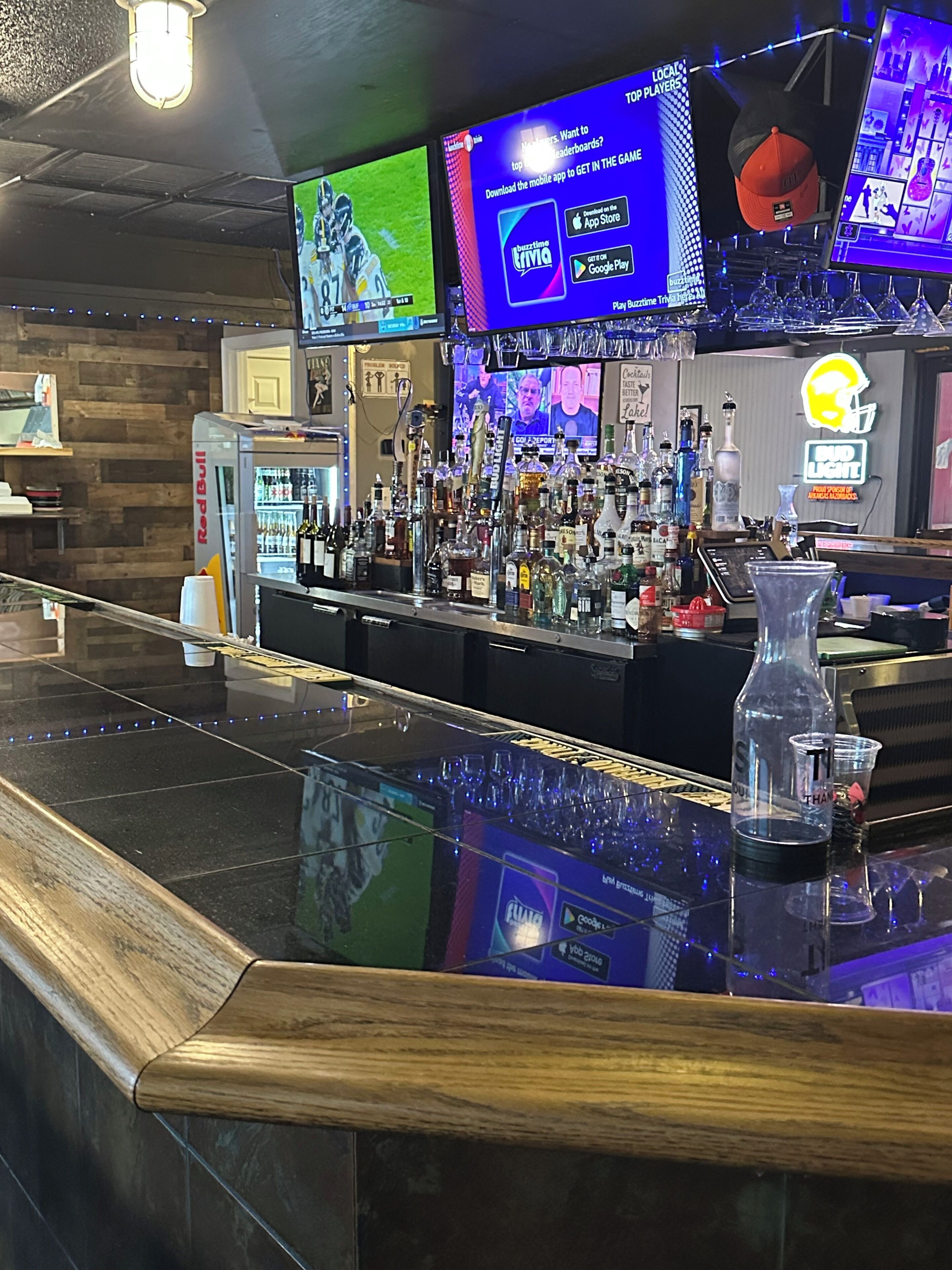 A bar area with multiple screens displaying sports and gaming content, a variety of liquor bottles, and a clear glass on the counter.