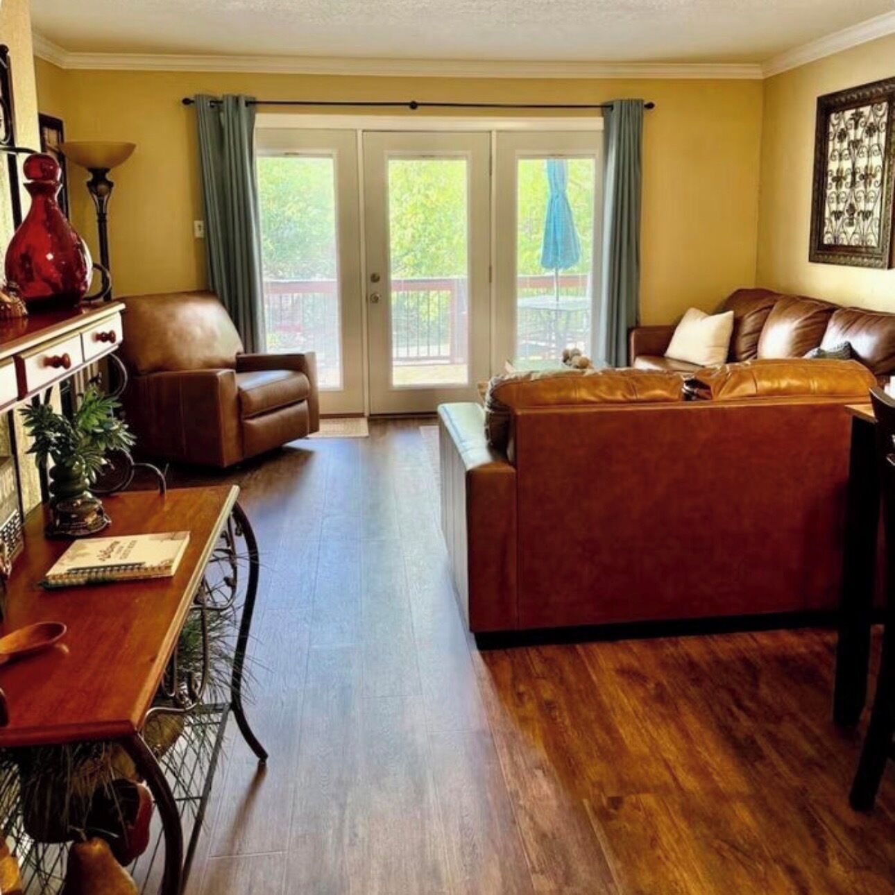 A living room with a brown leather sofa, a wooden dining table with chairs, and a sliding glass door leading to an outdoor area.