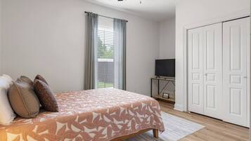 Ein Schlafzimmer mit einem Bett, einem Fernseher auf einem Schrank, einer weißen Tür und einem Fenster mit Vorhängen.