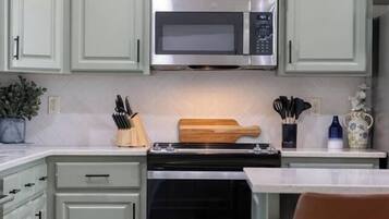 Une cuisine avec des meubles de couleur vert clair, un four à micro-ondes en acier inoxydable, un four noir et une planche à découper en bois.