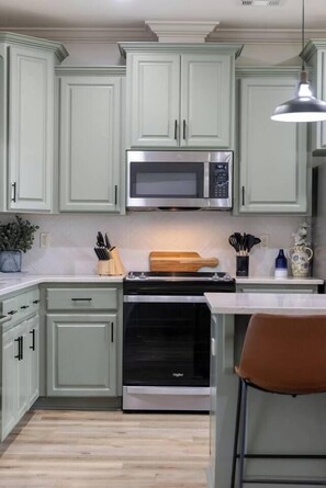 A kitchen with light green cabinets, a stainless steel microwave, a black oven, and a wooden cutting board.