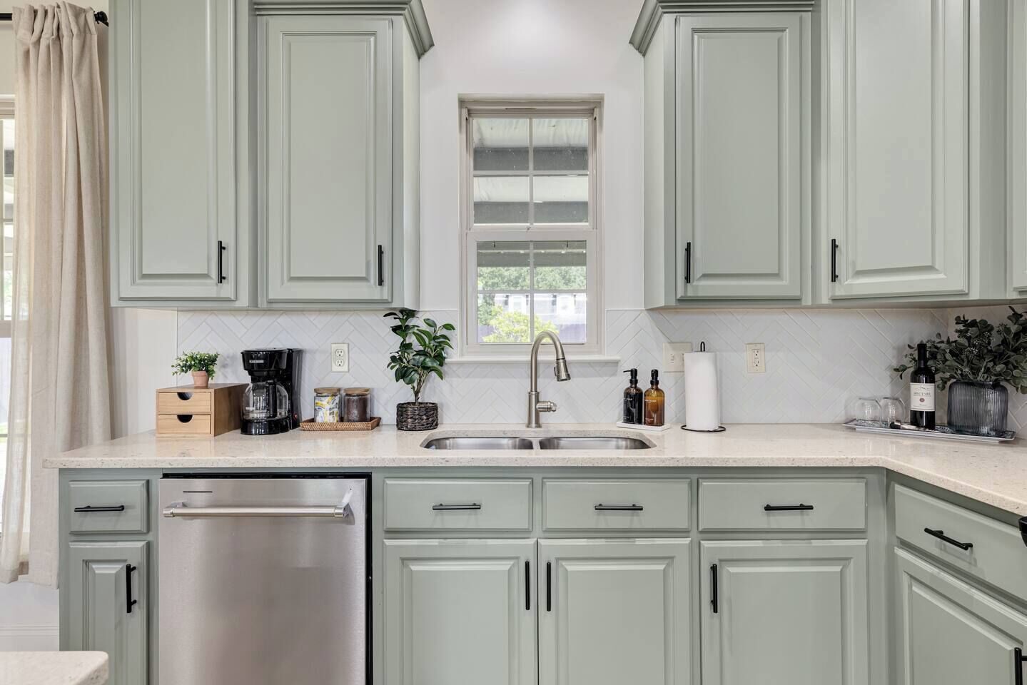 A kitchen with light green cabinets, a stainless steel dishwasher, a sink with a modern faucet, and various kitchen items on the countertop.