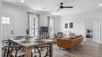 A modern living room with a sofa, a dining table with chairs, a television, and a ceiling fan.