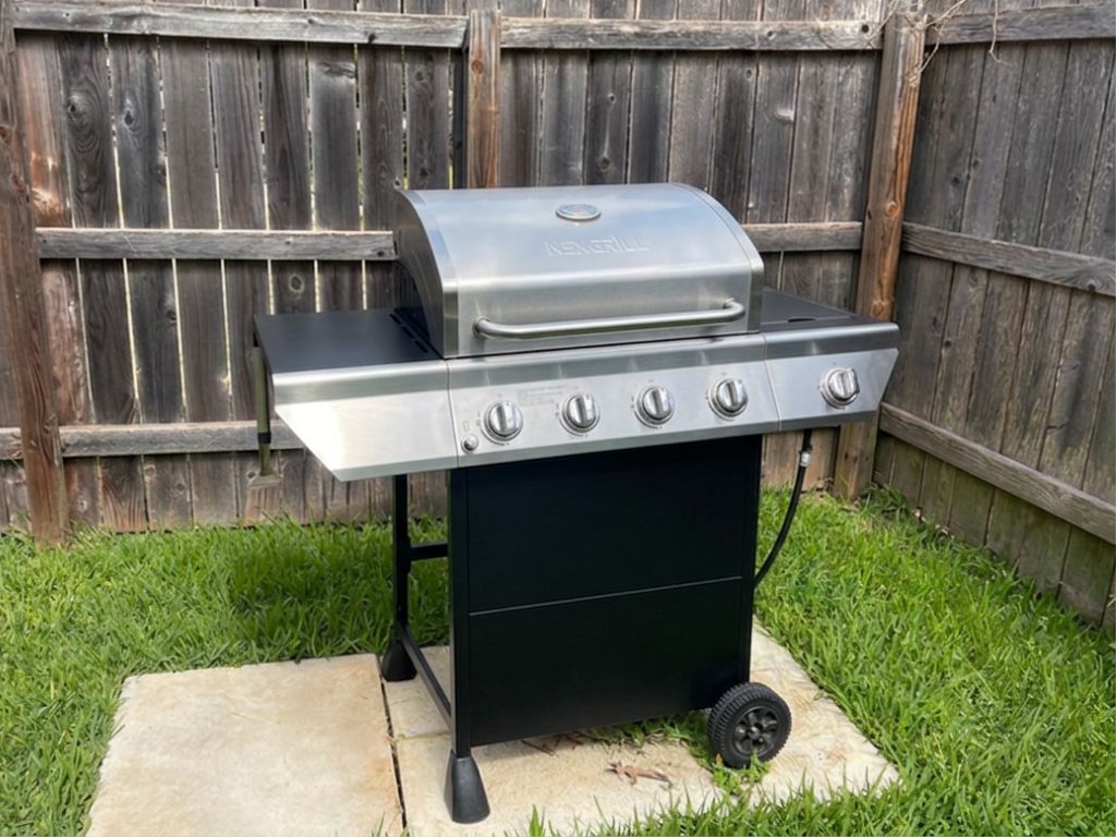 Una parrilla de gas portátil con cuatro quemadores y una repisa lateral, ubicada sobre una base de concreto en un patio trasero con césped y una valla de madera al fondo.