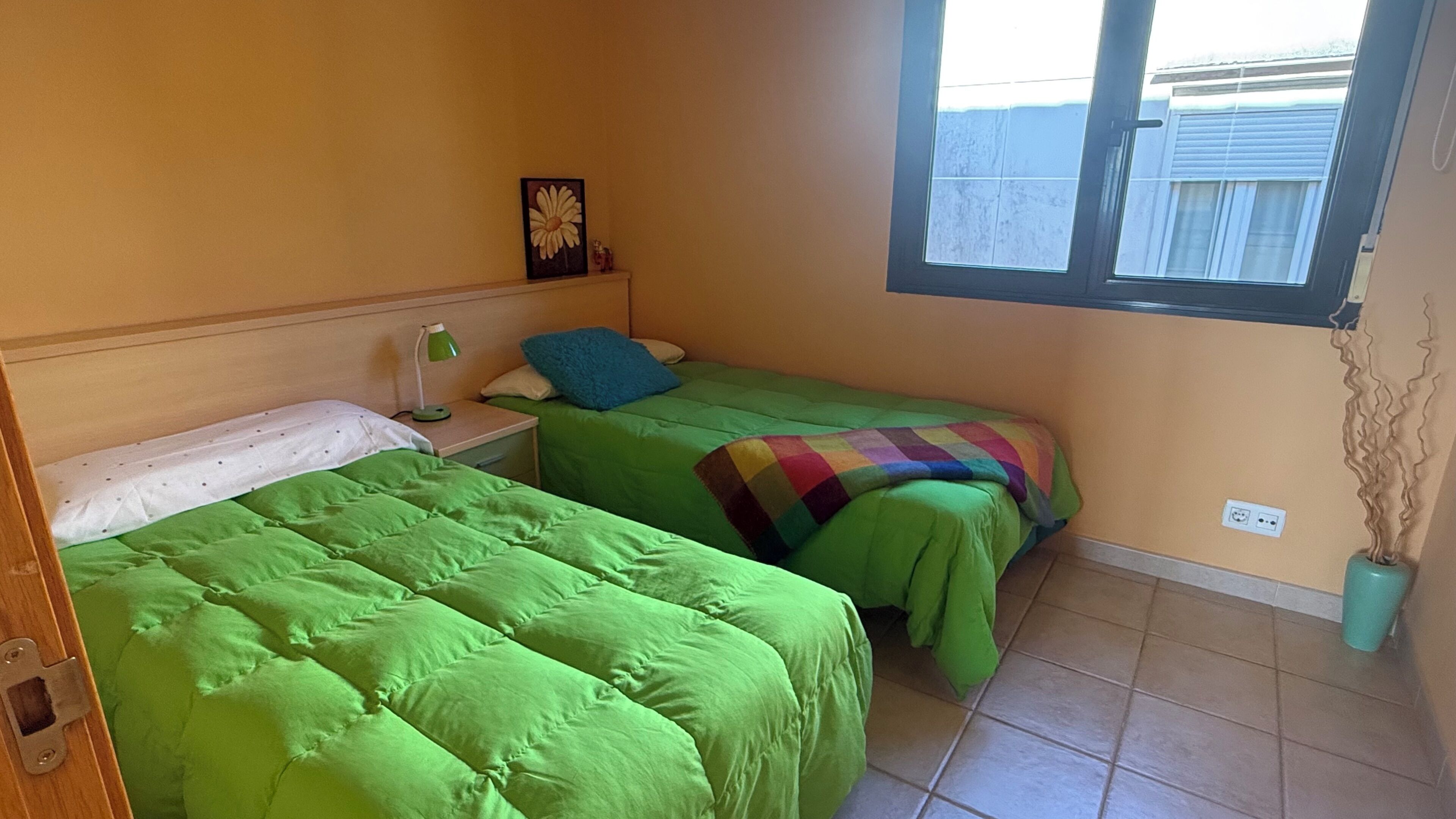 Una habitación con dos camas, una mesita de noche con lámpara, una ventana con persianas y un enchufe en la pared.