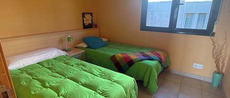 Ein Zimmer mit zwei Betten, einem Nachttisch mit Lampe, einem Fenster mit Jalousien und einer Steckdose.