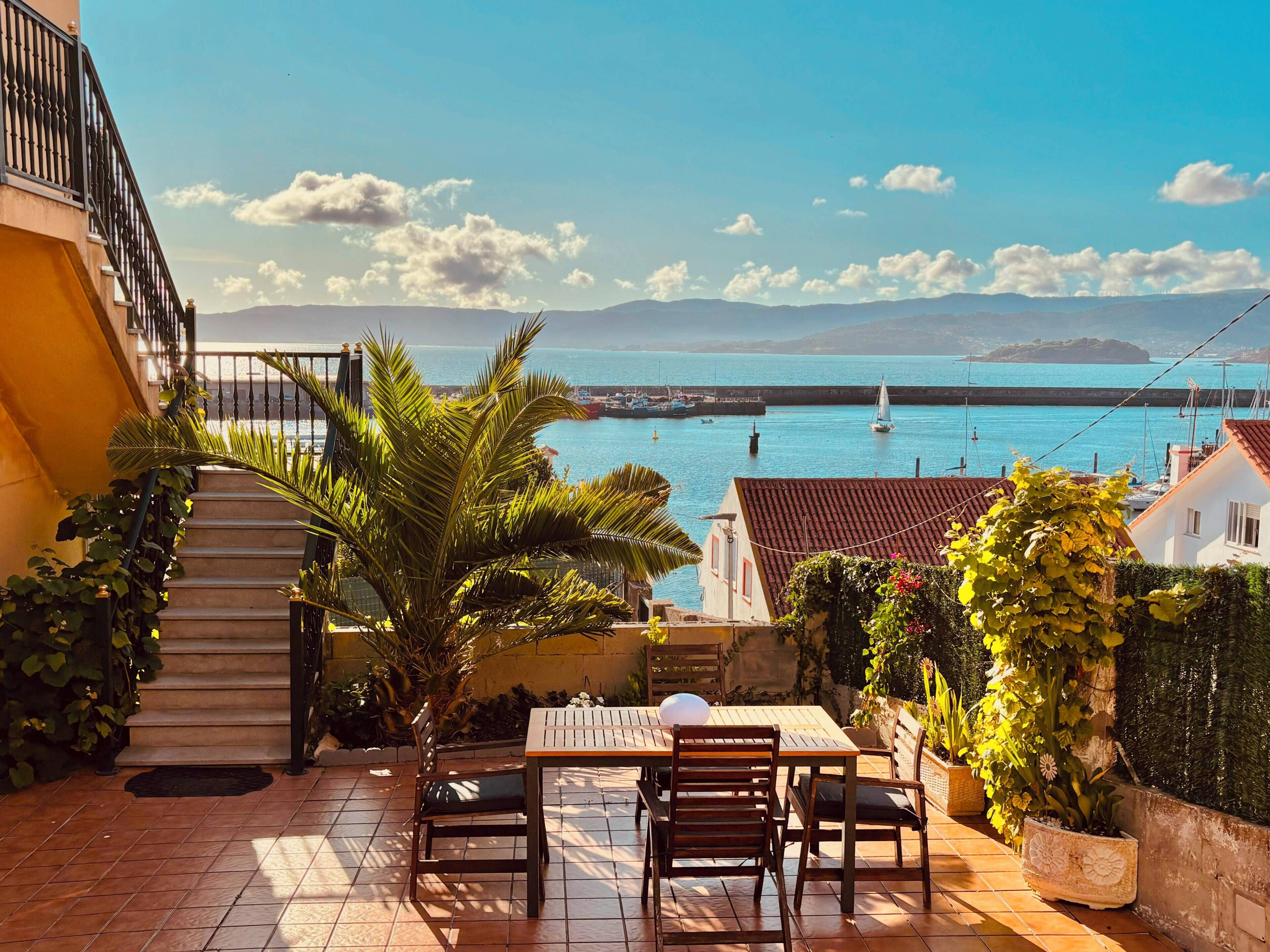 A patio with a table and chairs, overlooking a coastal view with boats and a clear sky.