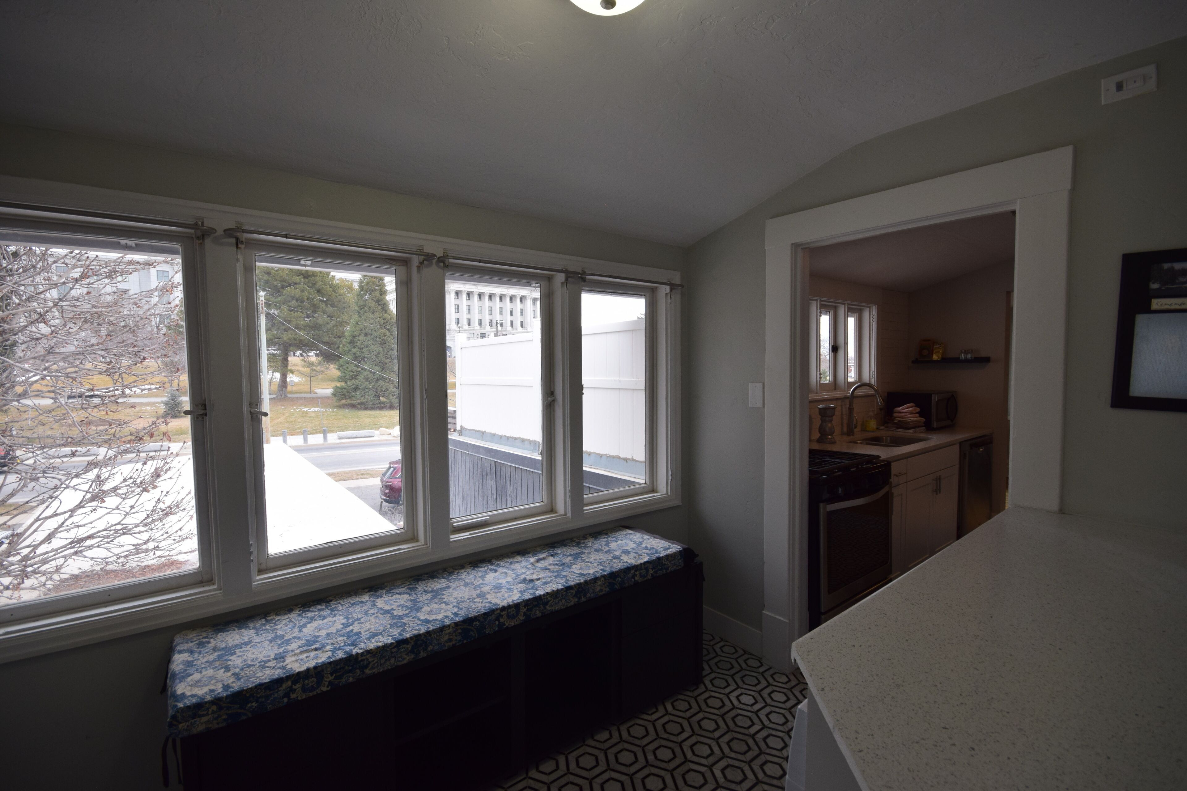 A room with a large window, a bench, a kitchen area, and a patterned floor.