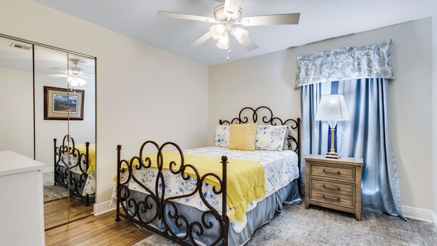 A bedroom with a bed, a nightstand, a mirror, a ceiling fan, and a window with curtains. - NEW! Kid Friendly 3BR Fenced Yard - Sports Complex (Youngsville)