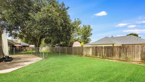 A backyard with a wooden fence, a grassy lawn, and a large tree. - NEW! Kid Friendly 3BR Fenced Yard - Sports Complex (Youngsville)