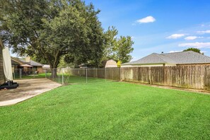 A backyard with a wooden fence, a grassy lawn, and a large tree. - NEW! Kid Friendly 3BR Fenced Yard - Sports Complex (Youngsville)
