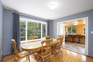A dining area with a wooden table and chairs, large windows, and a view of greenery outside.