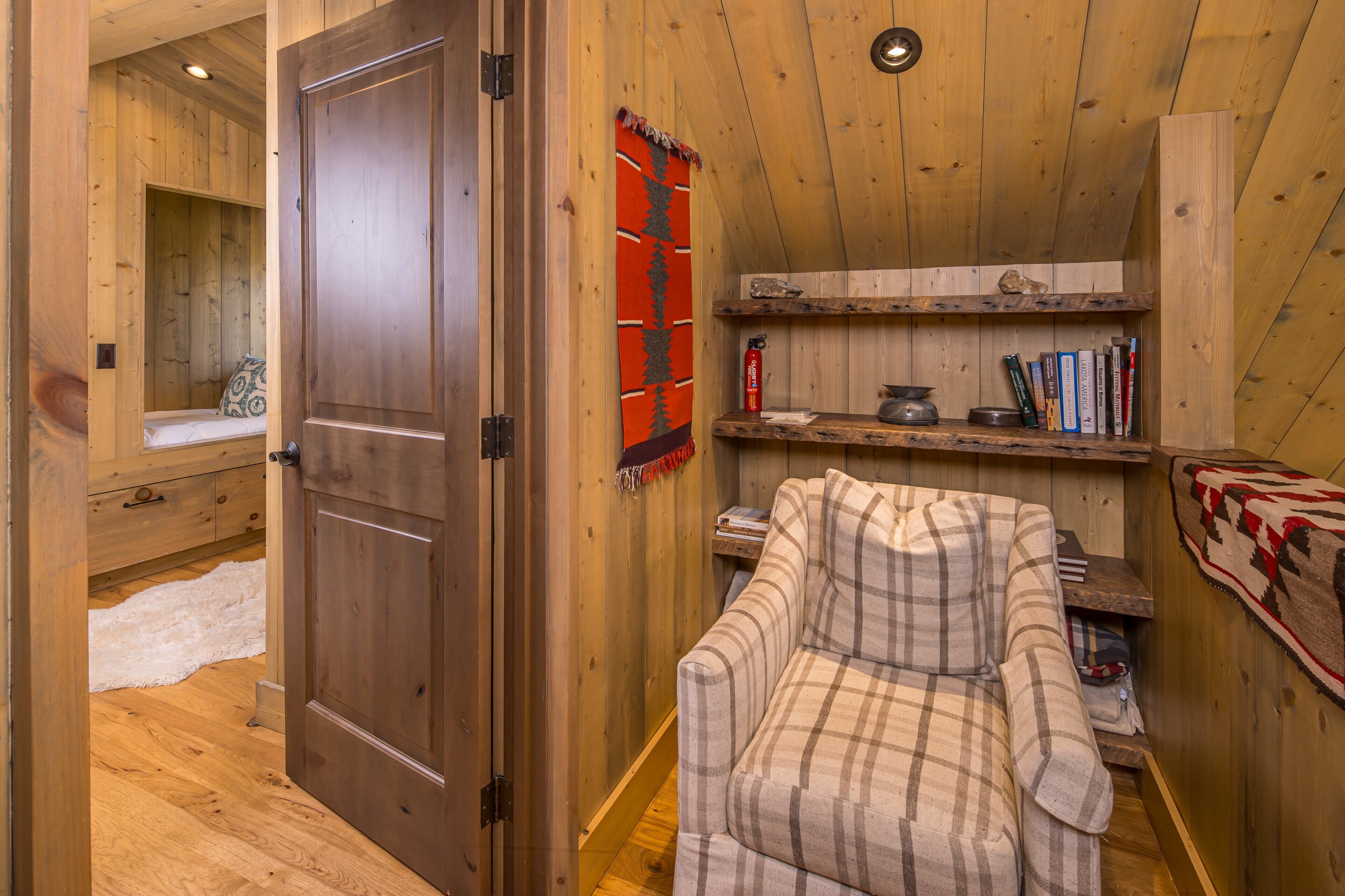 A wooden interior with a closed door, a plaid armchair, a shelf with books and a fire extinguisher, and a window with a view.