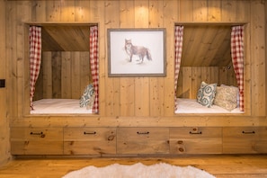 A wooden room with two beds, each with a checkered curtain, a framed picture of a fox, and a white fluffy rug.