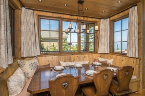A dining area with a wooden table set for six, large windows with curtains, and a view of greenery outside.