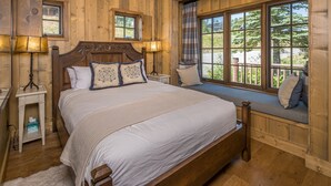 Un dormitorio de madera con una cama grande, dos lámparas de noche, un banco junto a la ventana con vistas, y una mesita de noche pequeña.