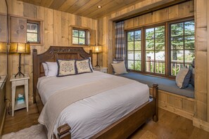 A wooden bedroom with a large bed, two bedside lamps, a window seat with a view, and a small bedside table. - NEW Listing | Ski In/Out + Private Hot Tub in Moonlight (Big Sky)