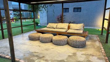 Uma área externa com assentos, incluindo um sofá e pufes, coberta por uma pérgola, e piso de concreto.