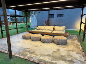 An outdoor seating area with a sofa and ottomans, a pergola overhead, and a concrete floor.
